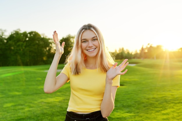 Gesturing young beautiful female teenager talking at camera student 16 17 years old giving interview looking at camera green sunset lawn in park background