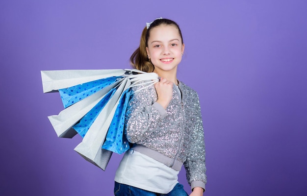 見栄えを良くします。ショッピングバッグの紫色の背景を持つ少女。ショッピングと購入。ブラックフライデー。セール割引。買い物の日。子供は束のパッケージを保持します。キッズファッション。誕生日の女の子。サプライズギフト。