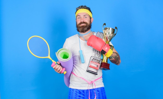 Get body ready for summer. Man bearded athlete hold sport equipment jump rope fitness mat boxing glove expander racket and golden goblet. Choose sport you like. Sport shop assortment. Sport concept.