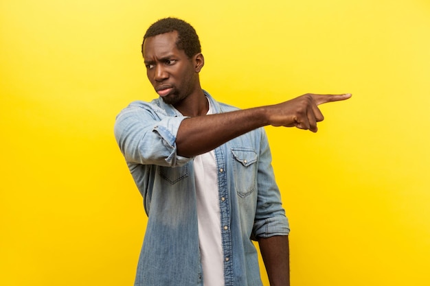 Get out. Portrait of irritated angry man in denim casual shirt pointing sideways, ordering to leave, looking with annoyed dissatisfied expression. indoor studio shot isolated on yellow background