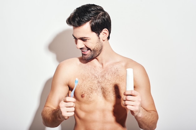 Get ready handsome darkhaired man holding toothpaste and toothbrush standing shirtless and smiling