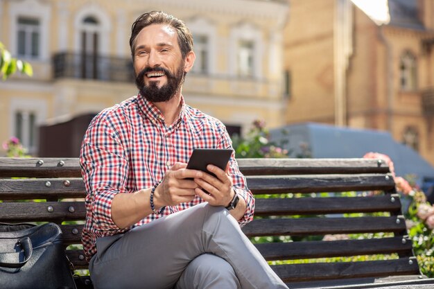 Get some rest. Joyful smiling bearded man sitting on the bench while holding modern gadget