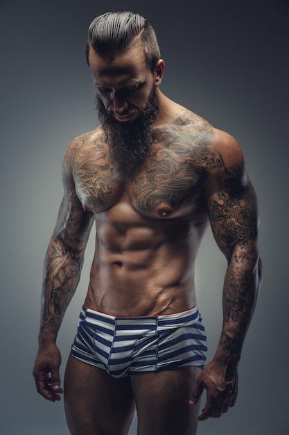 Getatoeëerde gespierde man met baard in strepen slipje. Geïsoleerd op grijze achtergrond.
