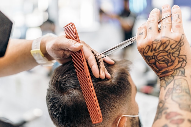 Getatoeëerde handen van een kapper die het haar knipt van een klant die een masker draagt in een kapperszaak