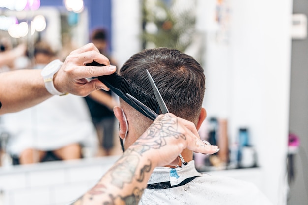 Getatoeëerde kapper die haar knippen van een blanke volwassen man met masker met een schaar en kam in een salon