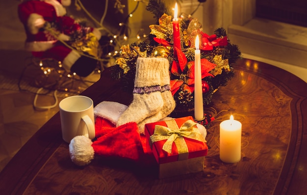 Getinte foto van kerstkaarsen, open geschenkdoos en wollen sokken op houten tafel bij open haard