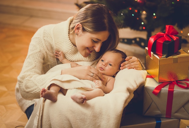 Getinte portret van jonge moeder die babyjongen kust, liggend in de woonkamer, versierd voor Kerstmis