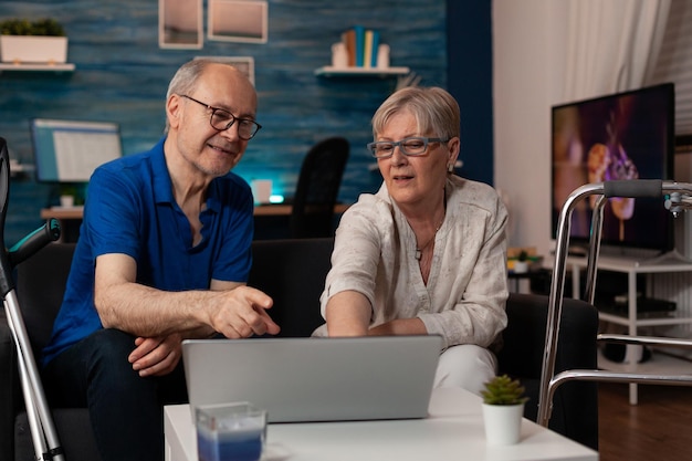 Getrouwd ouder stel dat samen naar het laptopscherm in de woonkamer kijkt. Gepensioneerde mensen die een apparaat gebruiken met technologie die thuis van vrije tijd genieten. Gepensioneerden met looprek en krukken