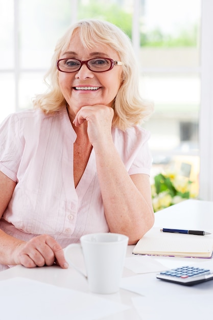 Getting finances straight. Happy senior woman holding hand on chin and smiling at camera while sitting at the table