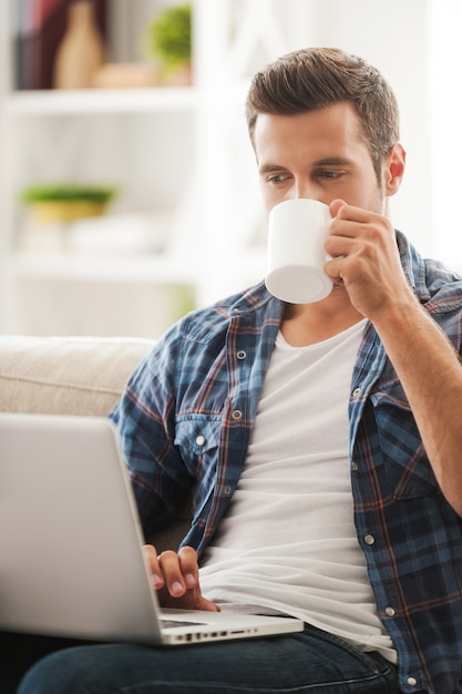 Ottenere le ultime notizie online. bel giovane che lavora al computer portatile e beve caffè mentre è seduto sul divano