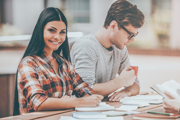 Getting ready for the next class. Smiling young woman writing in note pad and looking at camera while sitting with her friends at the wooden desk outdoors