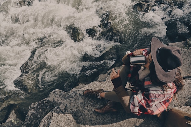 Getting ready to continue the journey. Top view of beautiful young woman covered with blanket drinking hot tee while sitting near the river in mountains