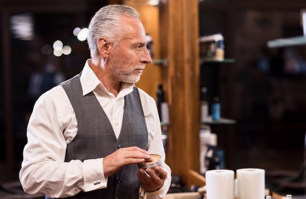 Getting ready. Side view of nice senior bearded man standing and holding jar with hair gel in front of mirror.