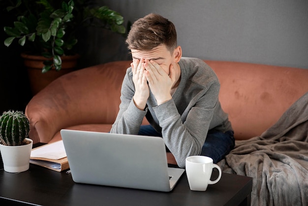 Foto gevoel van vermoeidheid frustreerde jonge man ziet er uitgeput uit en bedekt zijn gezicht met zijn handen terwijl hij aan zijn laptop zit thuis op zijn werkplek