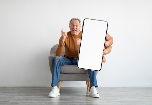 Geweldig aanbod opgewonden senior man zittend in een stoel en met lege smartphone