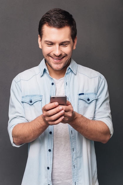 Geweldig bericht! Vrolijke jonge man die zijn smartphone met een glimlach gebruikt terwijl hij tegen een grijze achtergrond staat