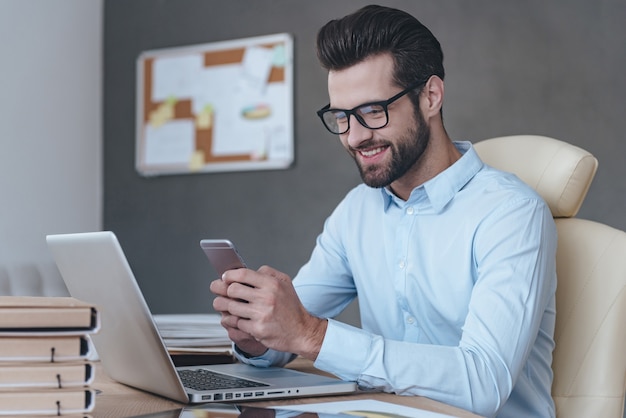 Geweldig nieuws. Knappe jonge man met een bril die een smartphone vasthoudt en ernaar kijkt met een glimlach