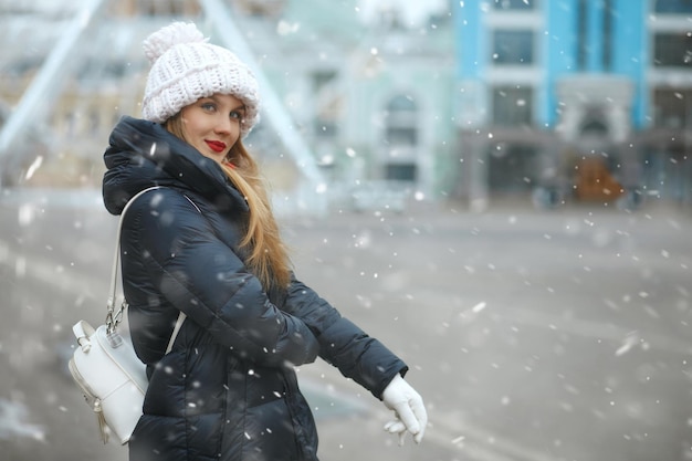 Geweldige blonde vrouw met een gebreide muts die tijdens sneeuwval in de stad loopt. Ruimte voor tekst