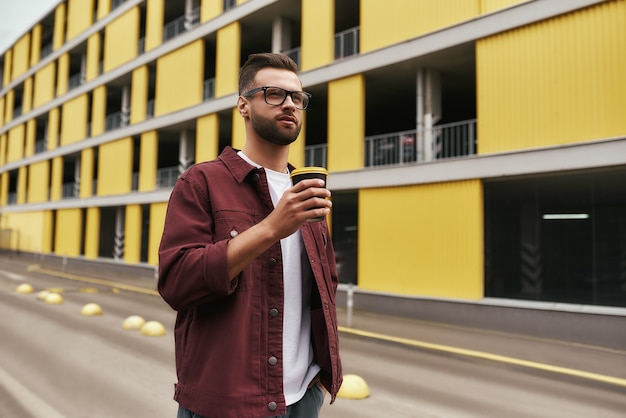 Geweldige dag knappe bebaarde man in vrijetijdskleding en bril met een wegwerpbeker terwijl