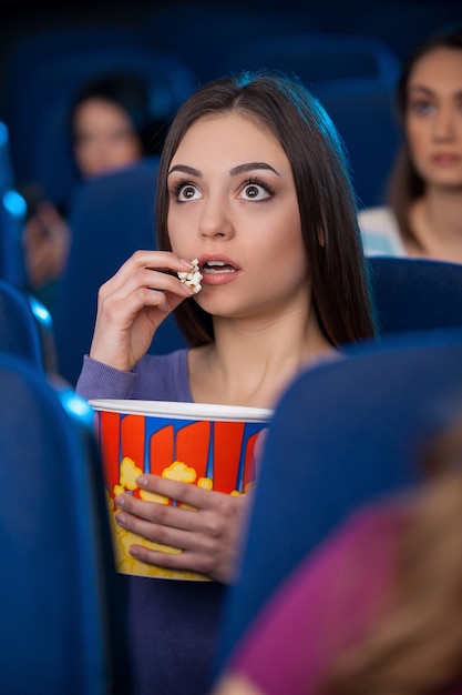 Geweldige film! Opgewonden jonge vrouw die popcorn eet en film kijkt terwijl ze in de bioscoop zit