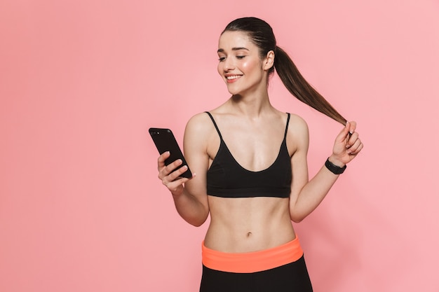 geweldige mooie jonge mooie fitness vrouw met behulp van mobiele telefoon geïsoleerd over roze muur