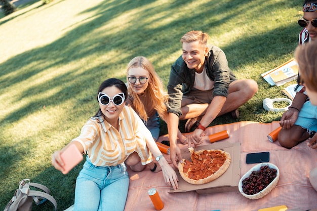 Geweldige tijd met vrienden. gheerful vrienden zittend op het gras selfie te nemen en pizza te eten na het doen van hun huiswerk.