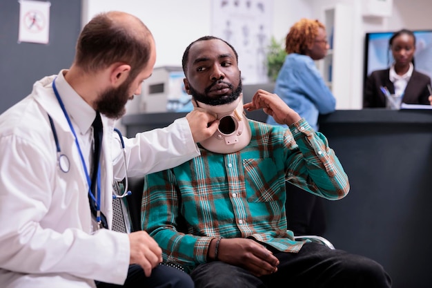 Gewonde Afro-Amerikaanse man met nekkraag in gesprek met arts op medische afspraak in wachtkamerlobby. Patiënt in pijn met cervicale schuimbrace na ongevalsverwonding, die behandeling krijgt.