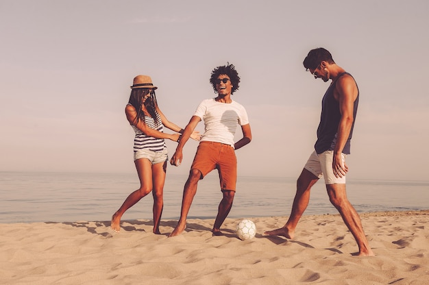 Gewoon lol hebben. Drie vrolijke jonge mensen spelen met voetbal op het strand met de zee op de achtergrond