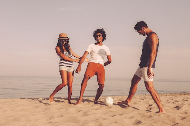 Gewoon lol hebben. drie vrolijke jonge mensen spelen met voetbal op het strand met de zee op de achtergrond