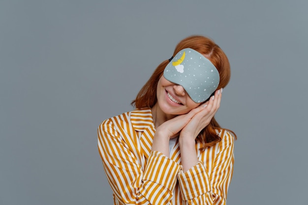 Foto gewoon ontspannen of tijd voor een dutje glimlachende zachte vrouw met oogmasker kantelt hoofd op geperste handpalmen draagt gestreepte pyjama poses over grijze muur kopie ruimte voor uw tekst voelt comfortabel en ontspannen