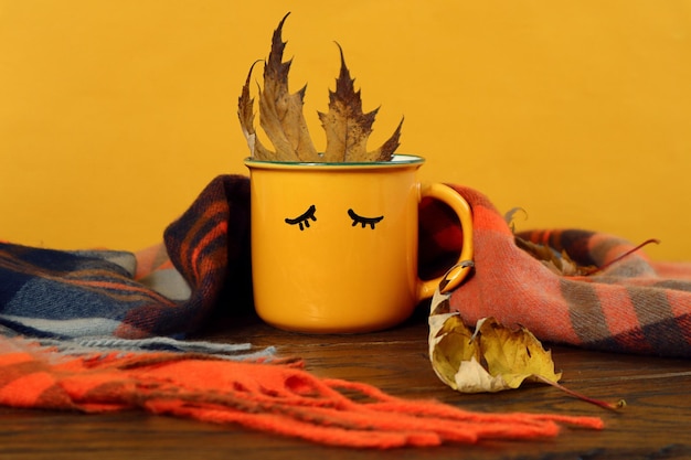 Gezellig herfst theekransje een gele mok met geschilderde slaapogen een esdoornblad erin bedekt met een geruite sjaal