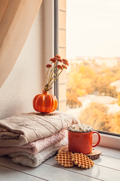 Gezellig herfststilleven op de vensterbank: warme wollen truien, pompoenen, esdoornbladeren en een kopje cacao met marshmallows en wafels.