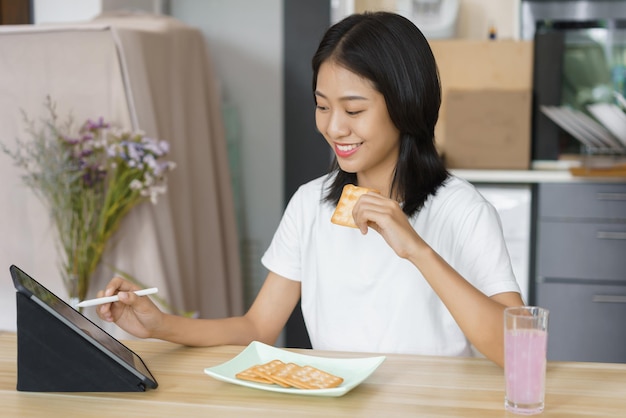 Gezellig levensstijlconcept jonge vrouw die tablet gebruikt en crackers eet terwijl ze thuis vrije tijd heeft