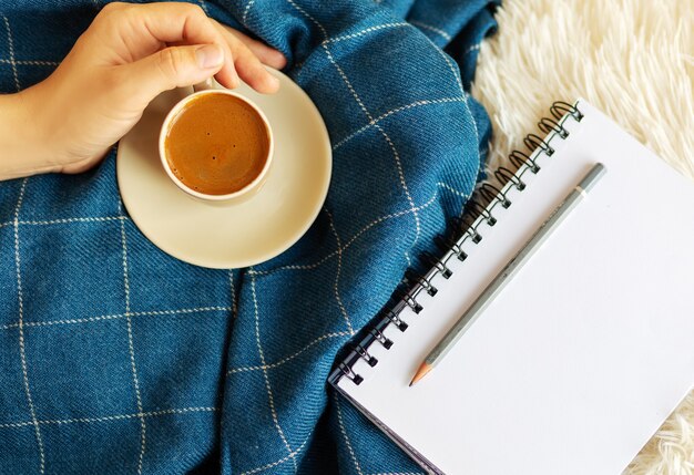 Gezellige flatlay met houten dienblad, handen houden kopje koffie, kaars, notitieboekje op witte pluizige lakens en blauwe deken. werk thuis concept. bovenaanzicht.