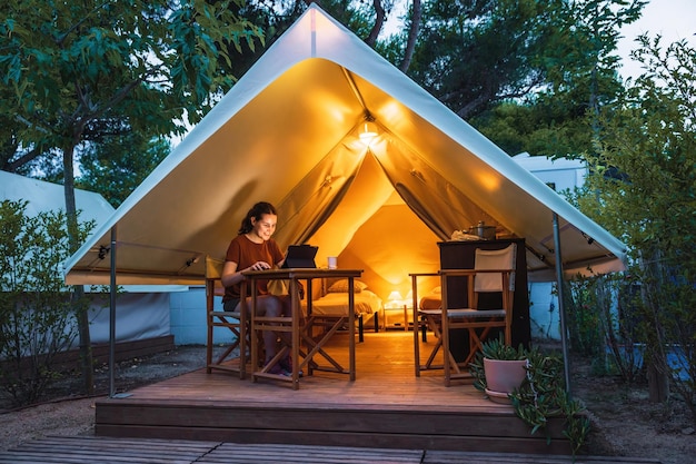 Gezellige open glamping-tent met licht erin en een vrouw die een laptop gebruikt tijdens luxe kamperen in de schemering