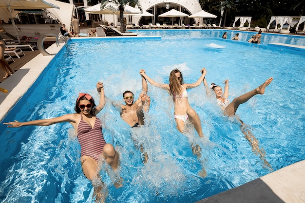 Gezelschap van jonge vrolijke meisjes en jongens springen samen hand in hand in het zwembad naast de loungezone in de open lucht op een zonnige zomerdag