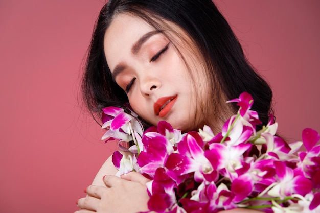 Gezicht geschoten Portret van Fashion 20s Aziatische vrouw mooi zwart haar express gevoel sensuele gelukkige glimlach. Meisje houdt paarse orchideebloem vast voor cosmetische huidverzorging en behandeling over roze achtergrond