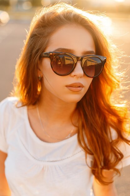 Gezicht portret van een mooie schattige jonge hipster roodharige vrouw in stijlvolle zonnebril in een trendy wit t-shirt bij een pretpark