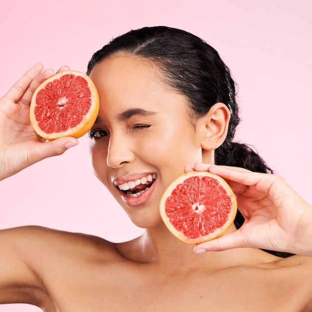 Gezicht schoonheid en vrouw knipoog met grapefruit in studio geïsoleerd op een roze achtergrond Portret natuurlijke cosmetica en model met fruit voedsel of voeding voor huidverzorging gezond veganistisch dieet of vitamine c