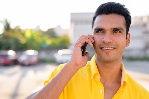 Gezicht van gelukkige jonge knappe Indiase man praten aan de telefoon buitenshuis