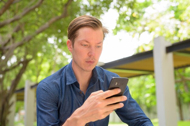 Gezicht van jonge knappe blonde zakenman met behulp van telefoon in het park buitenshuis