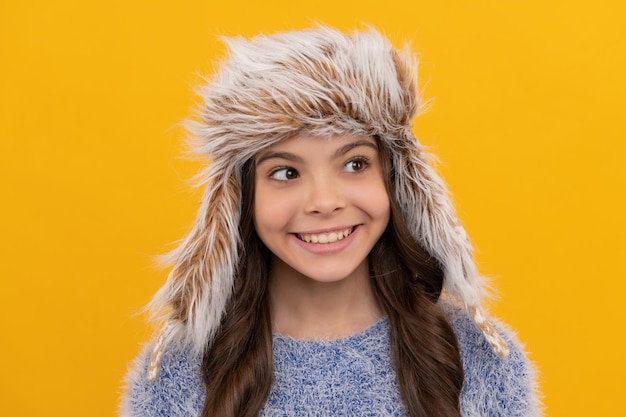 Gezicht van kind dragen warme kleding uitdrukken positieve emotie winter mode gelukkig kind met krullend haar in oorklep hoed vrouwelijke fashion model tiener meisje in gebreide kleding op gele achtergrond