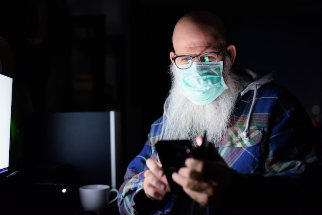 Gezicht van volwassen kale bebaarde man met masker met behulp van telefoon tijdens het werken vanuit huis 's avonds laat