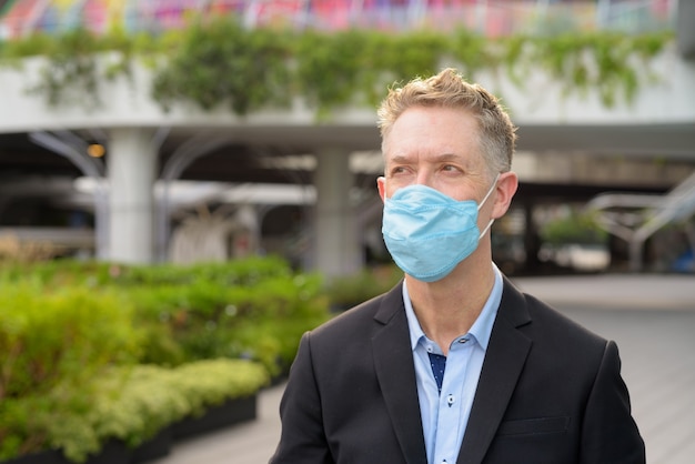 Gezicht van volwassen zakenman met masker voor bescherming tegen uitbraak van coronavirus, denken in de straten van de stad