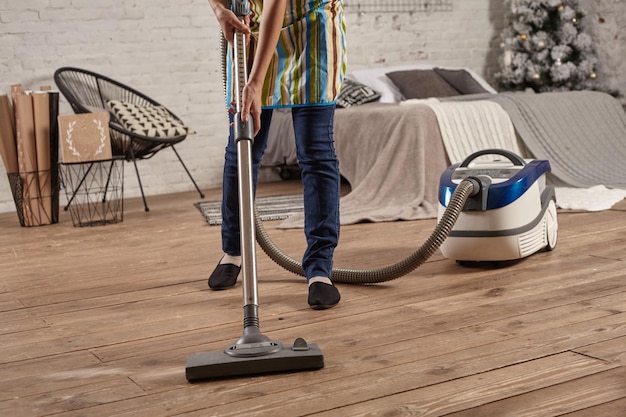 Gezichtsloos middengedeelte van een jonge vrouw die een stofzuiger gebruikt op de vloer van de huiskamer, schoonmaaktaken en klusjes doet, een nauwgezet interieur.