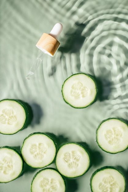 Gezichtspipet serum wei schoonheidsproduct in de buurt van plakjes komkommer in water spatten verse transparante vlekjes Verticaal