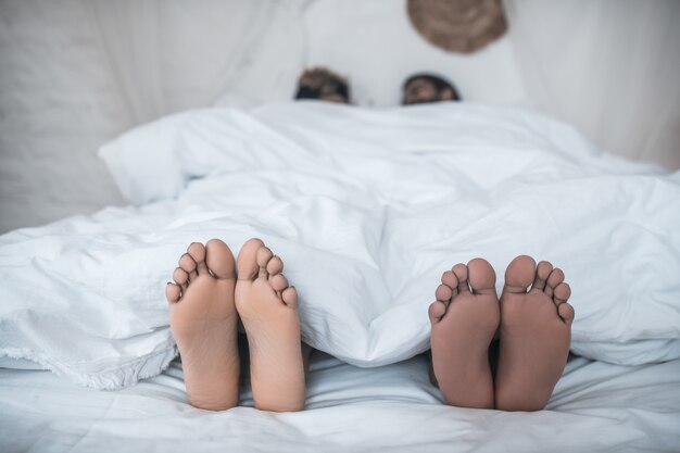 Gezinsgeluk. Voeten van donkere huid man en vrouw uitkijken onder de witte deken op bed thuis in de ochtend