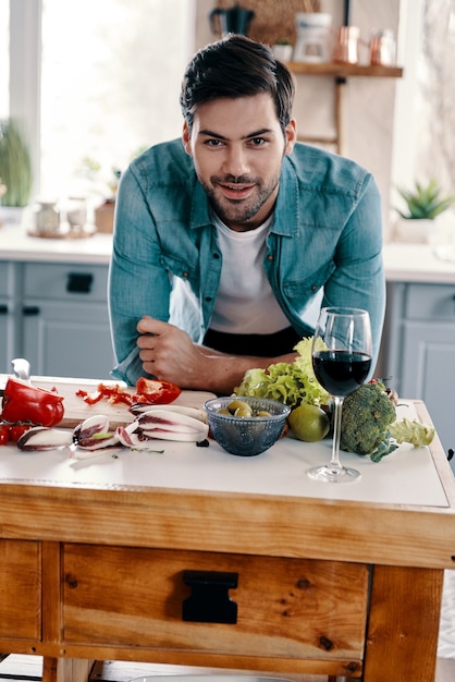 Gezond diner koken. knappe jongeman in vrijetijdskleding die naar de camera kijkt en glimlacht terwijl hij thuis in de keuken staat