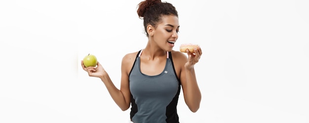 Gezond en dieetconcept Mooie sportieve Afro-Amerikaan maakt een beslissing tussen donut en groene appel Geïsoleerd op witte achtergrond