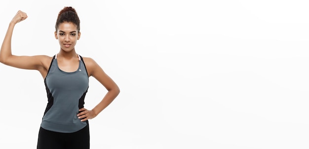 Gezond en Fitness concept Portret van jonge mooie Afrikaanse Amerikaan die haar sterke spier toont met zelfverzekerde vrolijke gezichtsuitdrukking Geïsoleerd op witte studio achtergrond